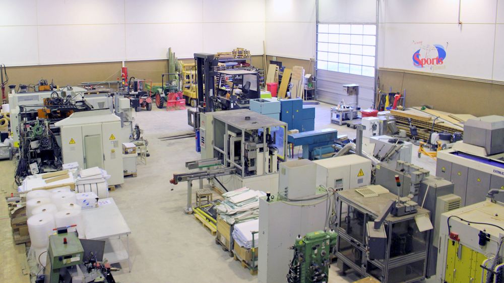 Weitblick in eine Fertigungshalle mit mehreren Maschinen, Materialbereichen, Rollenware, Stapler und Toranlage.